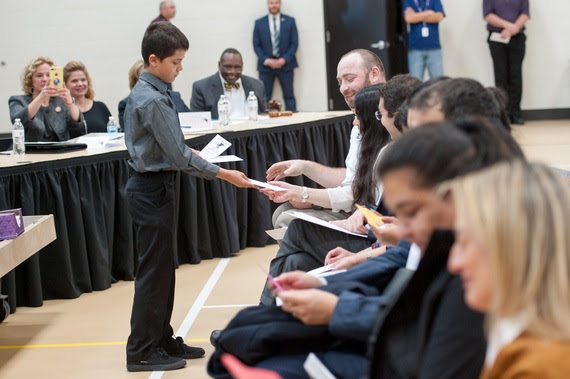 A fifth-grade boy hands cards to the citizens sitting in the front row of an auditorium.