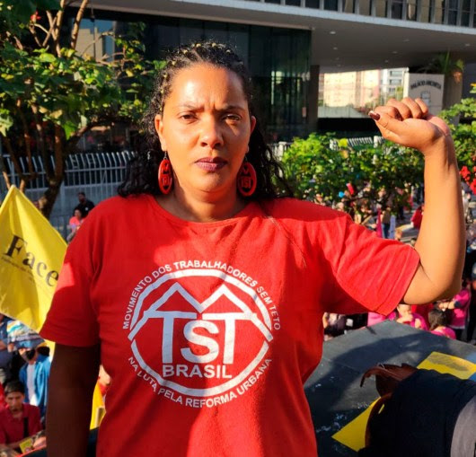 Débora Lima, coordenadora do MTST, durante manifestação contra o Plano Diretor em São Paulo. Débora é uma mulher negra, ela veste camiseta e brincos vermelhos com o logo do MTST e está com o punho cerrado em riste, em sinal de resistência