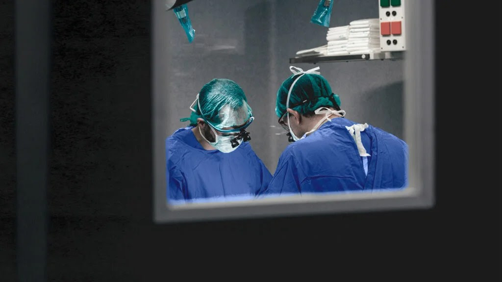 Two surgeons are seen operating on a patient through the window of an operating theater