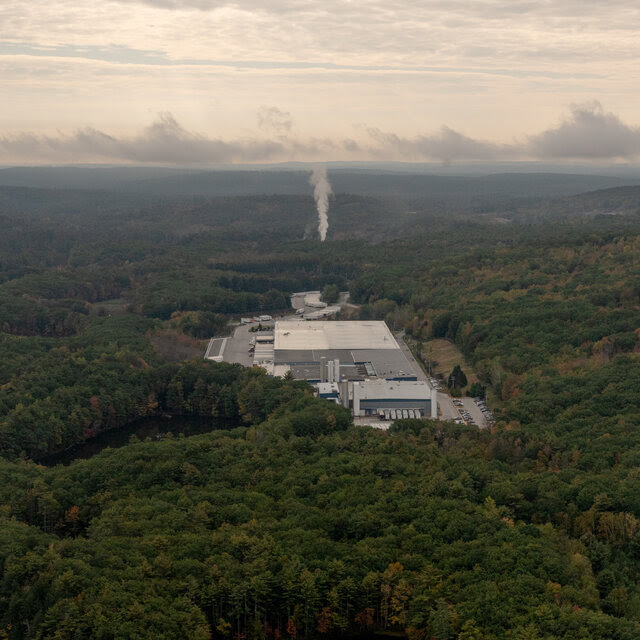 An aerial view of a large, gray industrial building nestled in an expanse of forest, with a body of water in the foreground.