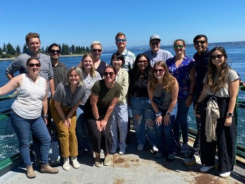 Fifteen people pose for a photo on the outdoor deck of a ferry on a sunny day