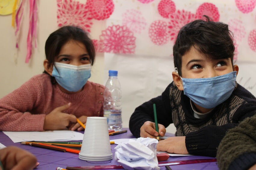 A young girl and young boy smile while colouring. Both are wearing masks.