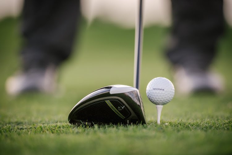 A golf ball sits in a tee on green grass, waiting to be hit by a golf club.