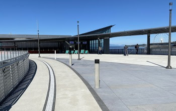Elevated pedestrian walkway with tactile pavement in some areas, views of the waterfront in the background and the ticket booth area in the distance 