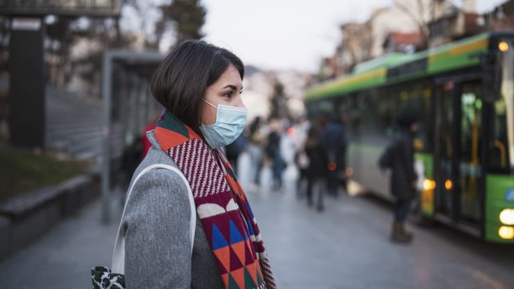 a woman waits for a bus wearing a medical mask