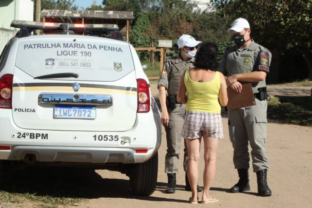 Ao lado de uma viatura da Patrulha Maria da Penha, dois policiais
militares (uma mulher e um homem) conversam com uma mulher que aparece de
costas em uma rua de chão batido.