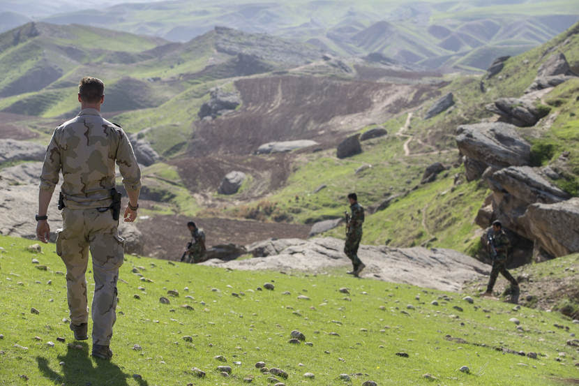 Nederlandse militaire trainer kijkt in groen berglandschap toe hoe Iraakse militairen oefenen.