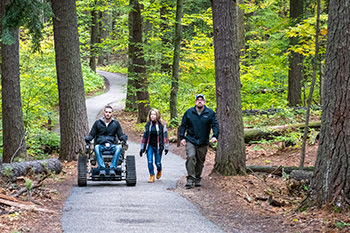 visitor using track chair and two other visitors on wooded trail