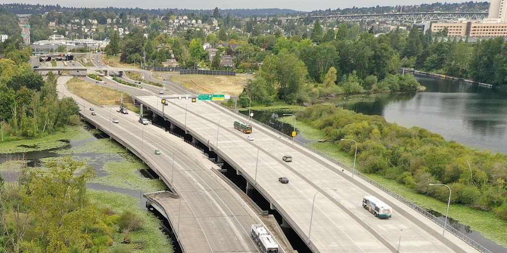 An aerial view of a highway bridge over a body of water in front of a residential neighborhood. There are multiple buses and cars on the highway.