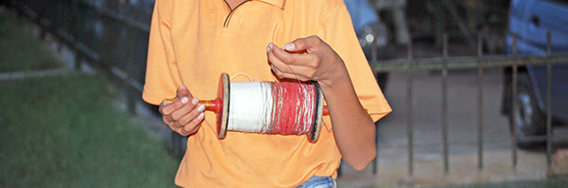A picture of a young boy in an orange shirt holding a spindle of red string while is left hand grasps a section of the thin red string.