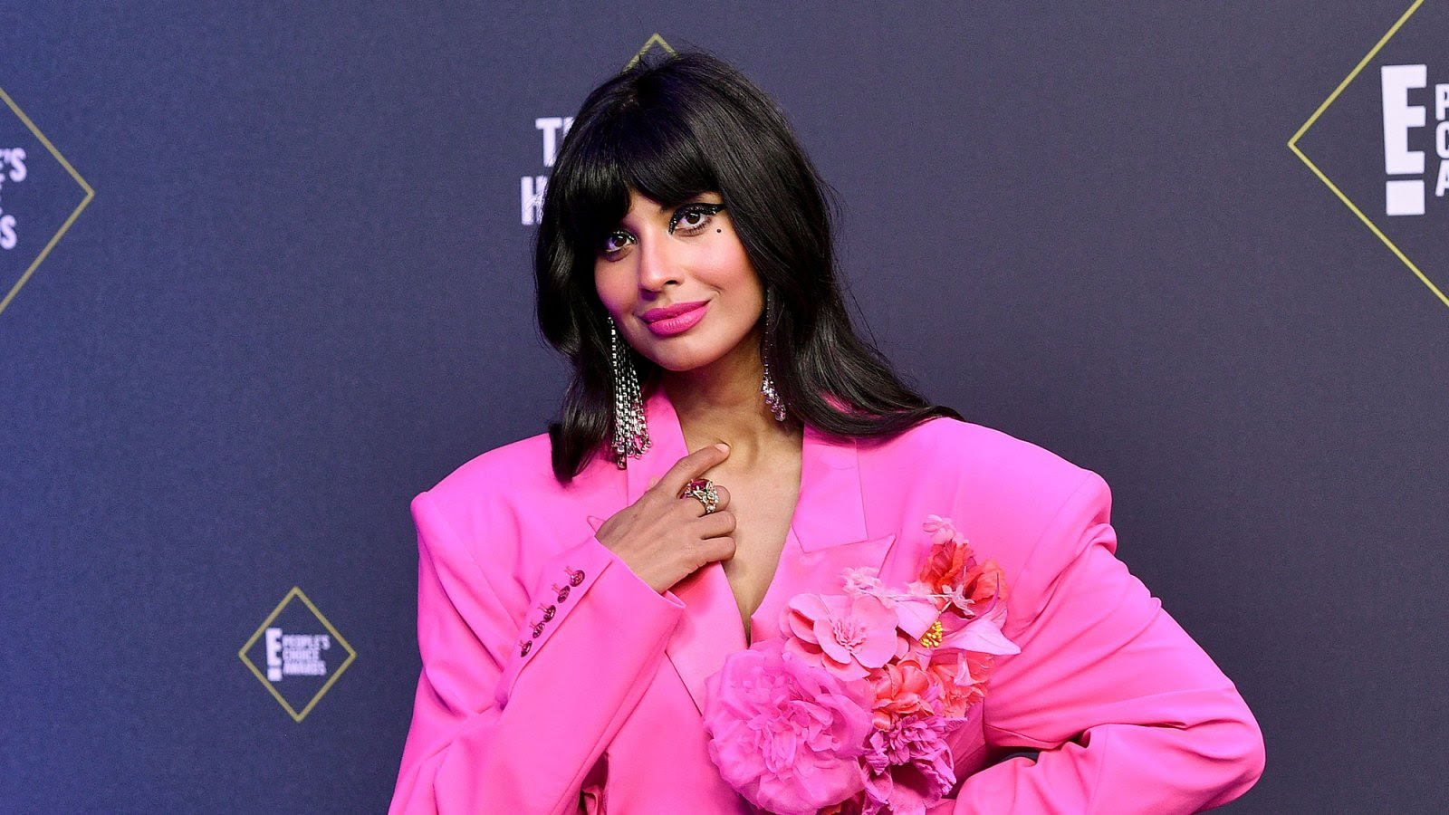 Jameela, a woman with long black hair, is standing in front
of a black wall. She wears a bright pink outfit with pink flowers and pink lipgloss. 
