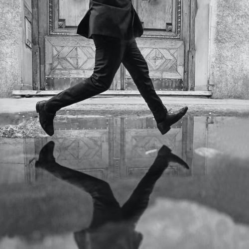 black and white image of man stepping over a puddle that he's reflected in