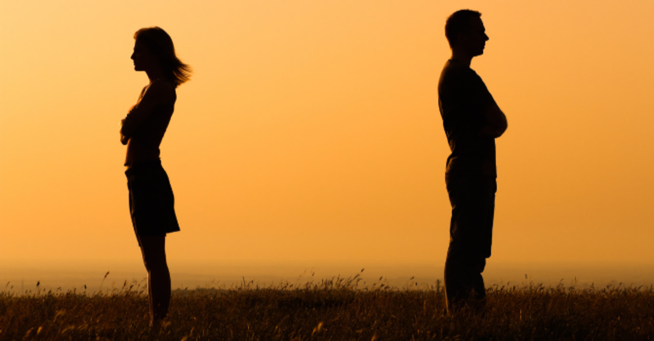 Couple standing apart with their backs to each other and arms crossed are silhouetted against the sunset