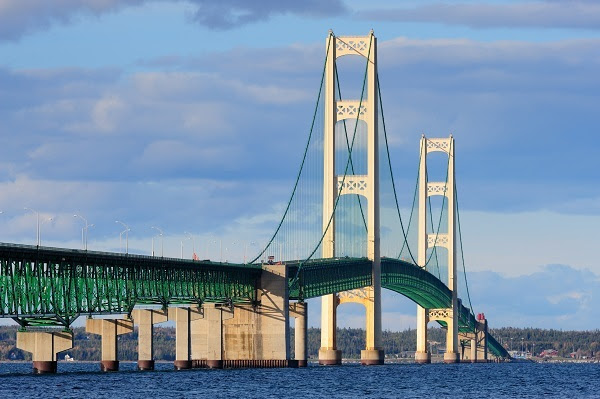 long view of a huge, beige and pale green, gently arced suspension bridge over deep blue water, and blue sky and pale clouds behind