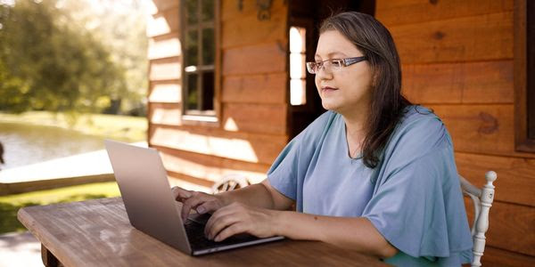 A white woman on a lap top sitting outside a remote cabin