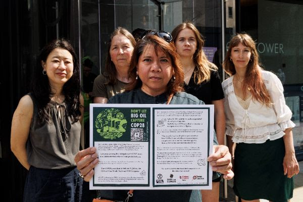 Five people standing in front of a building. The person at the front holds out a leaflet about concerns that COP28 is being coopted by the fossil fuel industry