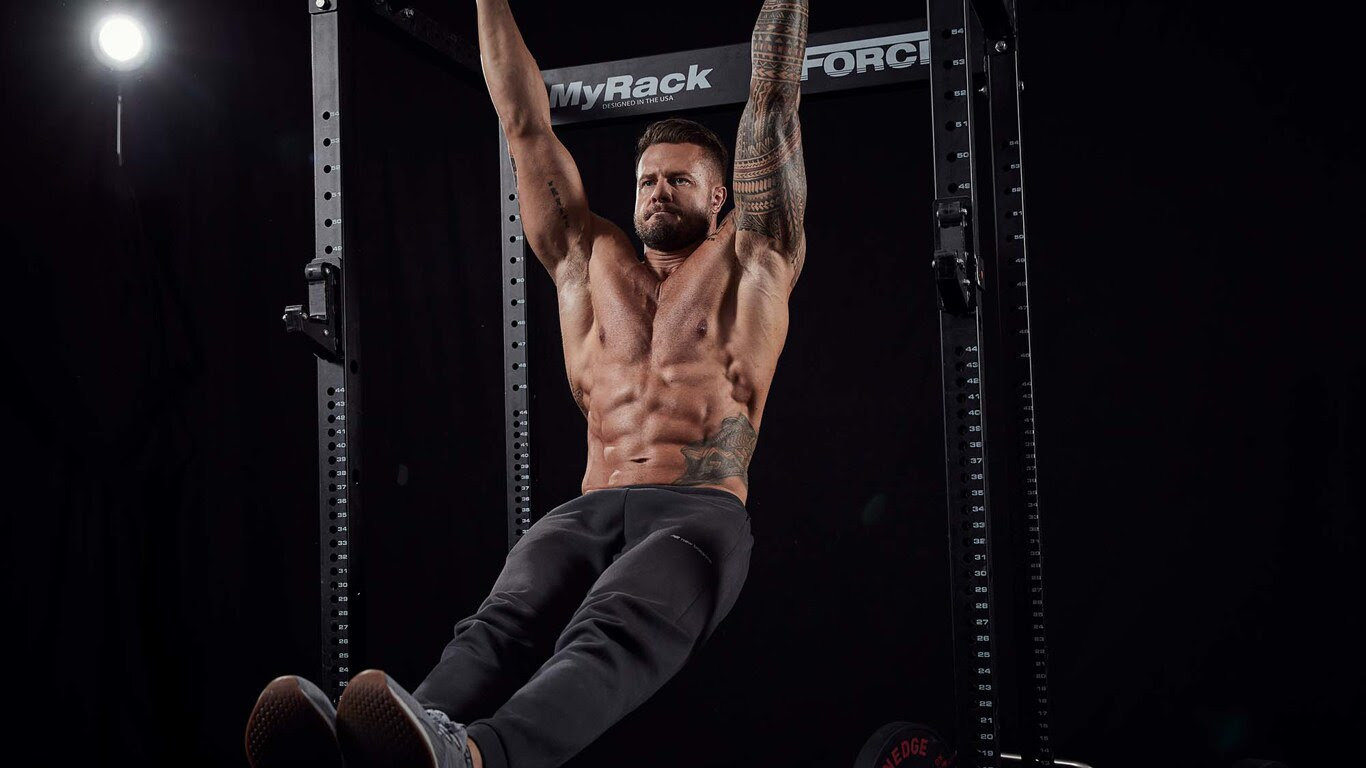 A shirtless, muscular man with visible abs hanging from a pull-up bar.