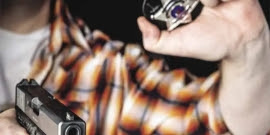 A man in a plaid shirt holds a police badge in one hand as he points a gun at the viewer.