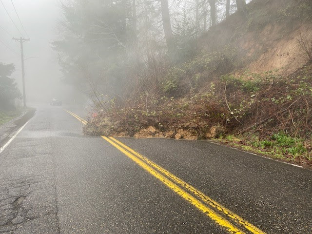 A landslide covers half of two-lane road, with debris from the adjacent hillside reaching the center double yellow line.