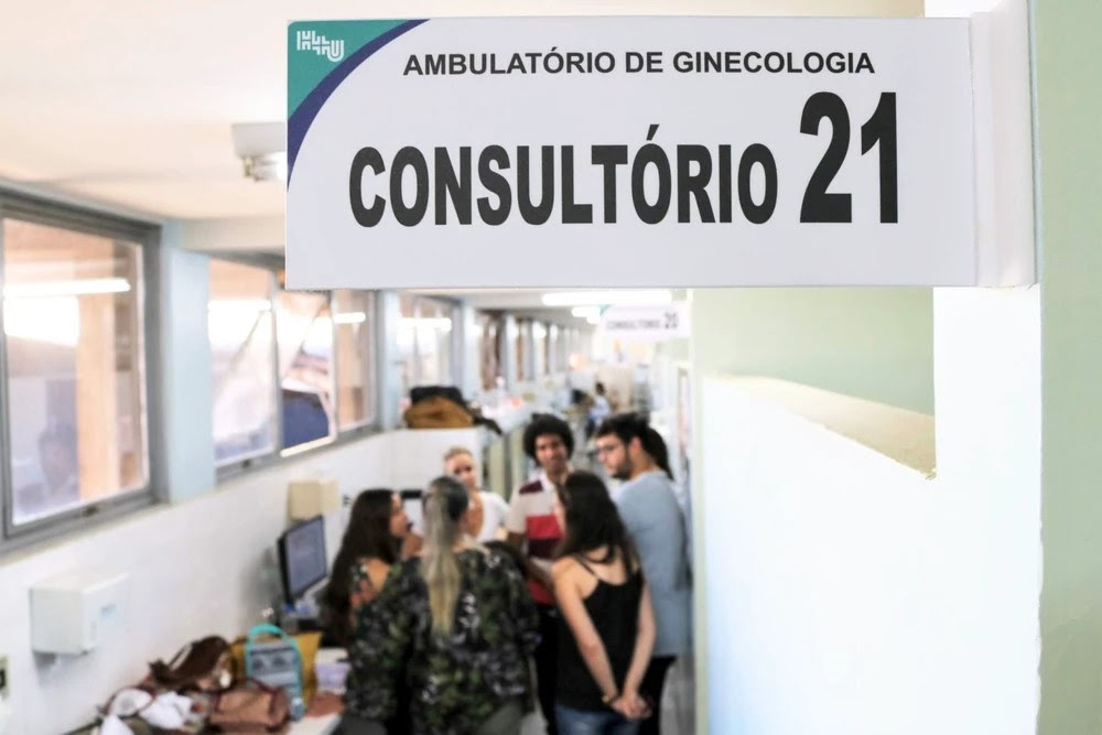 Imagem com foco em placa sinalizadora do Hospital das Clínicas de Uberlândia, escrito "Ambulatório de ginecologia - consultório 21". Em segundo plano há funcionários do hospital conversando