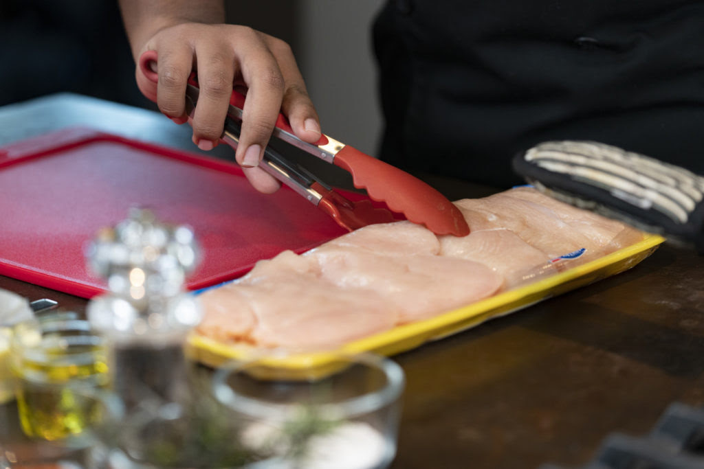 A hand gripping a pair of tongs takes raw chicken breasts out of packaging 