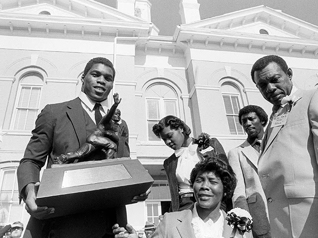 University of Georgia's Heisman Trophy winner Herschel Walker holds up his trophy for the crowd to see in front of the Johnson County courthouse in Wrightsville, Ga., Dec. 18, 1982. Several thousand fans, friends and well wishers turned out to honor Walker at the appreciation day at his hometown. Walker's mother Christine is in the center and his father Willis is standing at right. Others are unidentified. (AP Photo/Ric Feld)