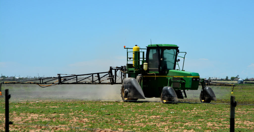 A green tractor with an attached arm spraying for pests is in a field. It illustrates the importance of drift management.