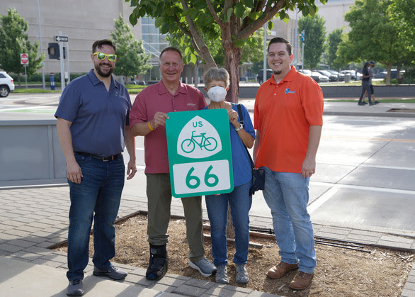 Photo of four people standing in front of a tree outdoors and holding up a road sign for U.S. Bicycle Route 66
