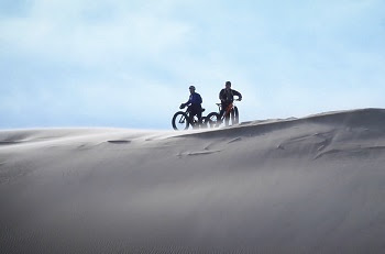 two people on fat-tire bikes are outlined in shadow, sitting atop tall sand dunes as sand swirls around them, backed by blue sky and clouds
