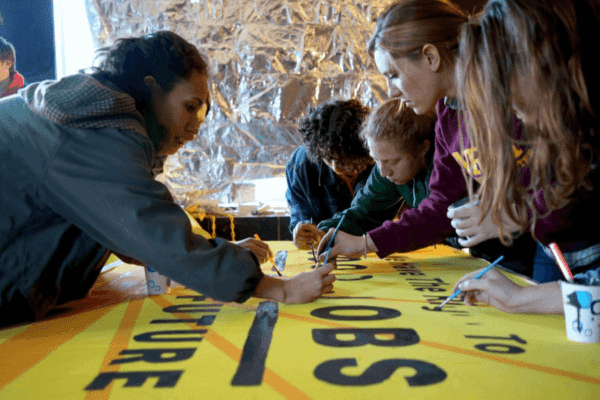 Art build for Sunrise Movement action in Washington DC, unknown photographer. 5 young people circle around a bright yellow sign and are drawing or painting various elements on it. The sign contains a slogan including the words "good" "jobs" and "future."
