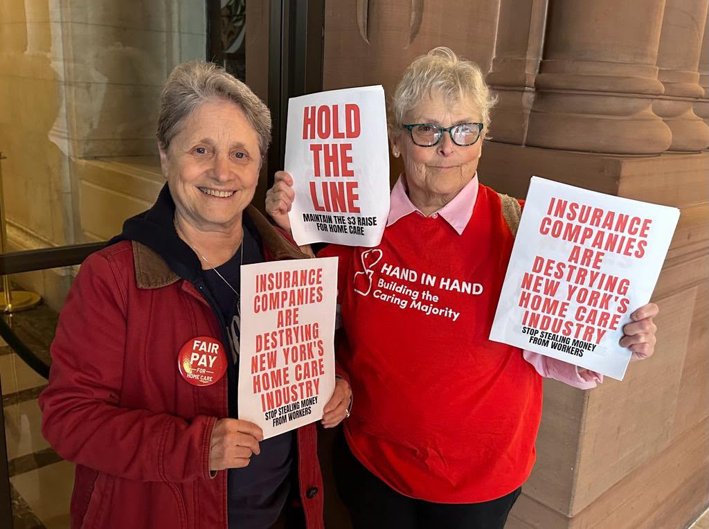 Photo of two older women dressed in red, holding signs that say ''hold the line'' and ''insurance companies are destroying New York's home care industry''