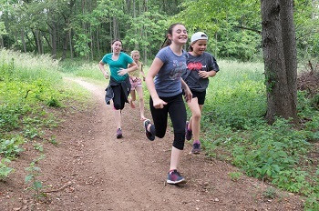 small group of girls and boys in T-shirts, shorts and jeans smile as they run down a dirt trail lined with green trees