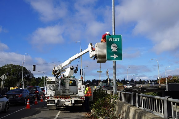 A construction worker in a lift installs a directional sign on a pole on the side of a road.