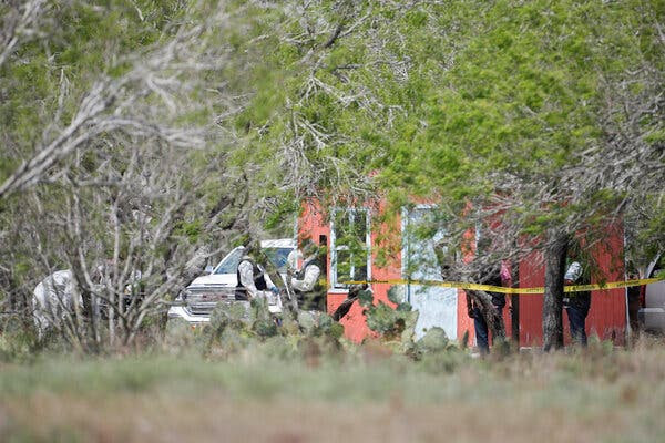 Through a thicket of trees, officials can be seen outside a small house with crime-scene tape visible.