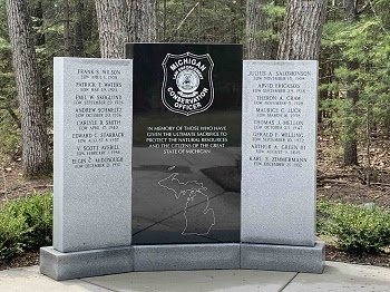 a gray-and-black, square granite memorial with officer names etched and the DNR logo, surrounded by tall, green trees