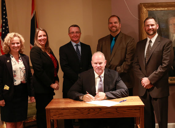Governor Mead sits at a desk to sign the proclamation with State Superintendent Jillian Balow and four principals standing behind him.