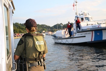 back of a woman conservation officer in uniform on a DNR boat, with white and red U.S. Coast Guard vessel and two people in the background