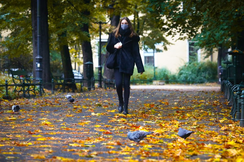 A young woman wears a face mask while walking through a park with autumn leaves.
