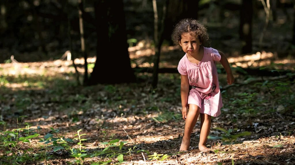 A young girl in the woods scratching her leg due to a bug bite.