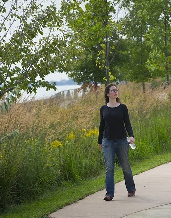 A dark-haired woman in a black shirt and light jeans, holding a water bottle, walks along a paved trail in a wooded area near a river