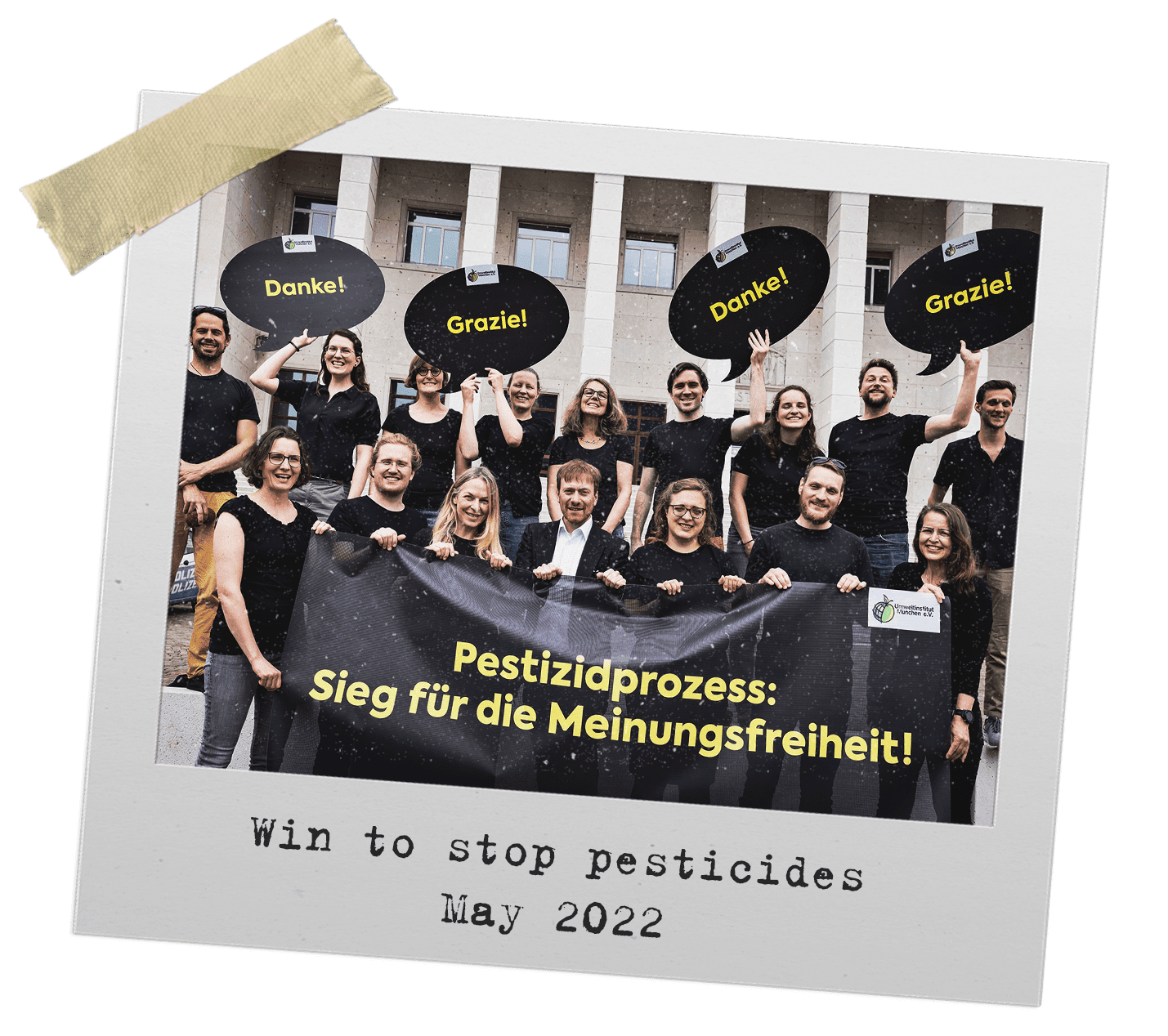 Two rows of people wearing black t shirts stand in front of building. Front row (7 people) hold banner, back row (9) people hold speech bubble-shaped signs saying Thank You in different languages.