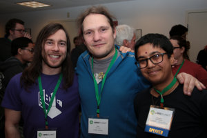 Friends enjoying the pre-conference open house. Their arms are around each other and they are smiling, wearing LibrePlanet nametags.