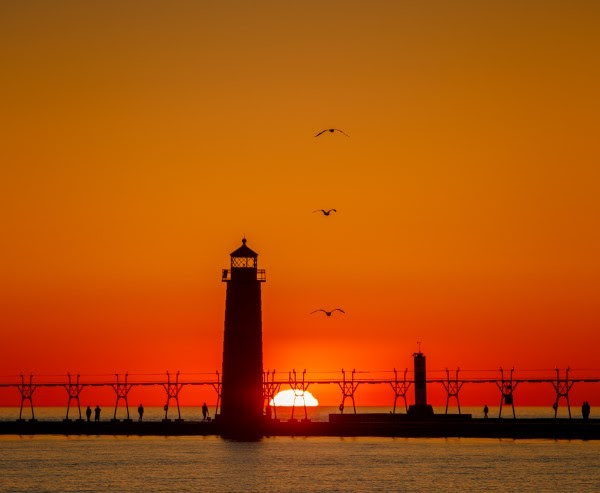 A lighthouse and pier are backlit by the gradient red and oranges of a brilliant sunset as the shadows of three gulls fly overhead.