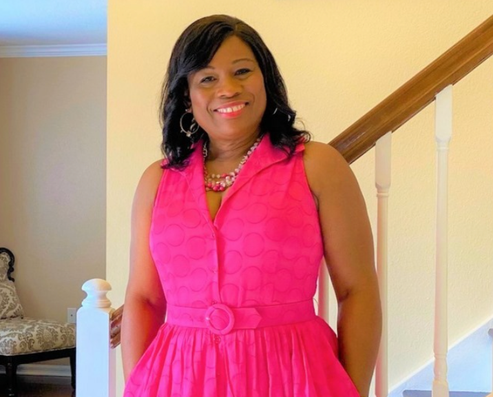 A black woman with curly black hair and a pink dress. She is smiling