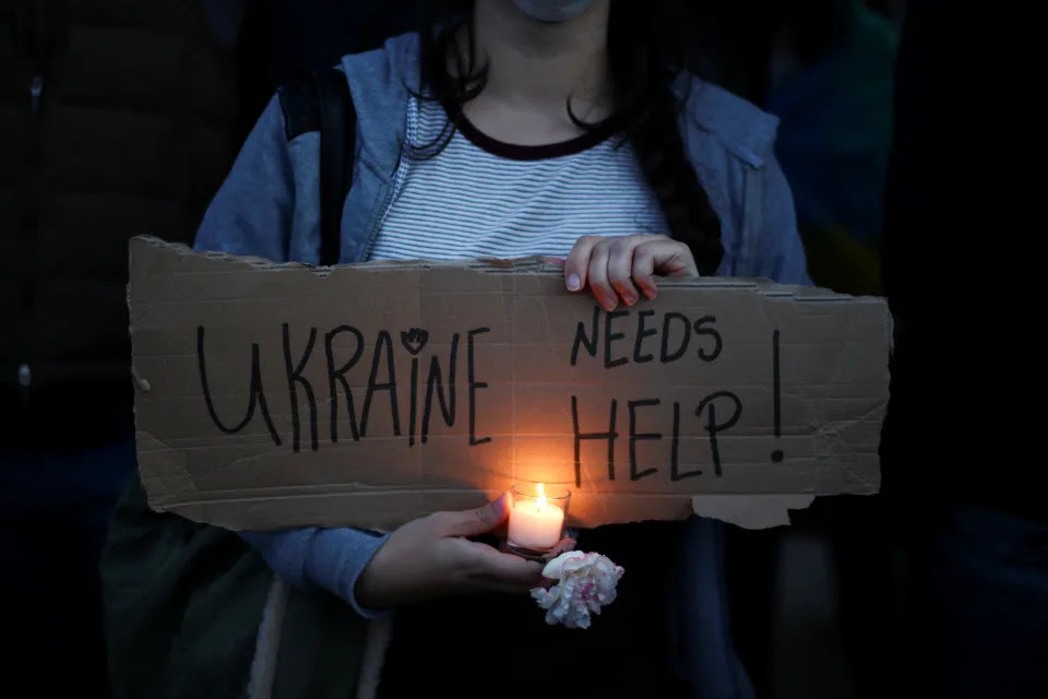 A person holds a candle and a sign reading Ukraine Needs Help! at an antiwar protest.