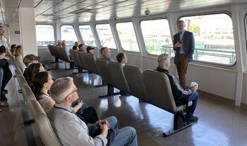 Person standing in front of ferry passenger cabin speaking to several people sitting in chairs