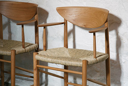 A photograph of two wooden, rush seated chairs desisned by Honours Furniture. They are displayed against a textured white wall, one is fully visible and the other is only half in the frame.