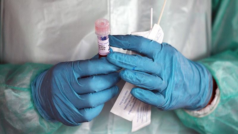 Two hands in blue gloves holding testing tube with lid.