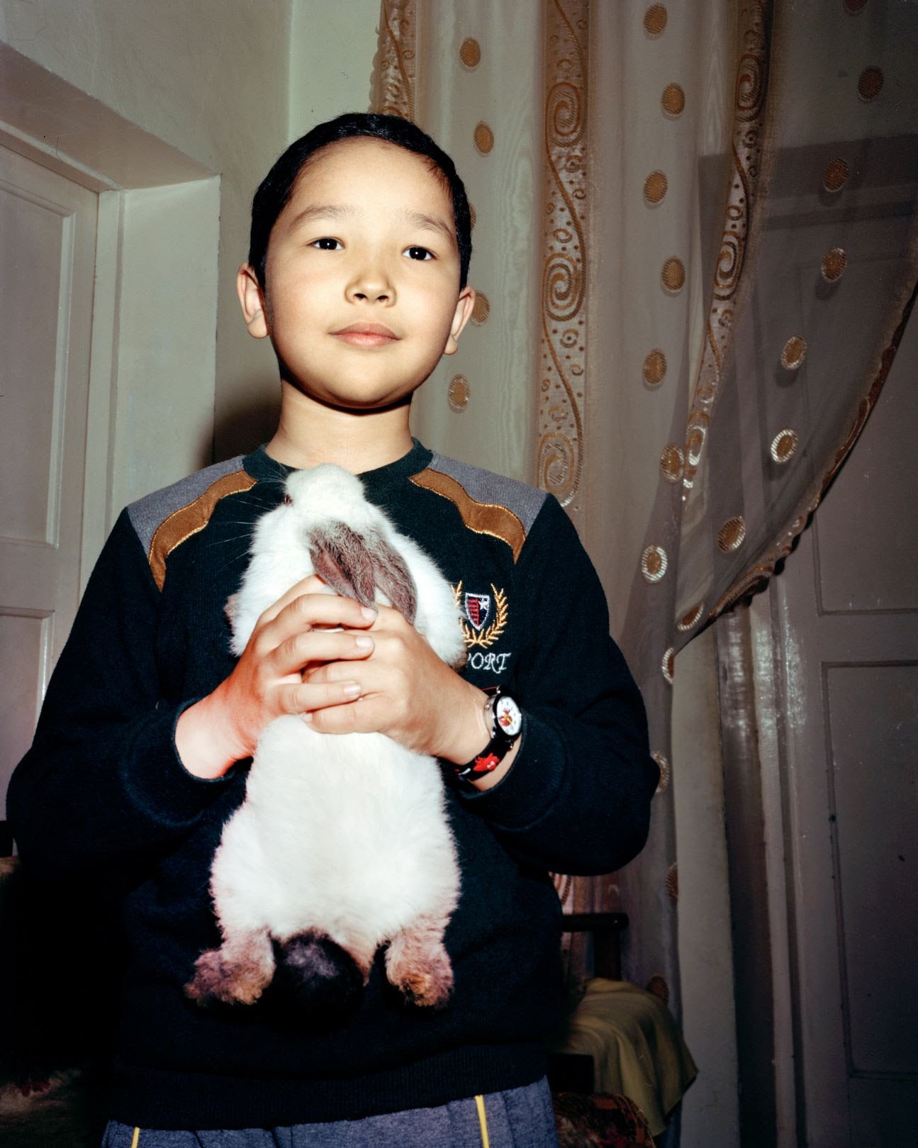[IMAGE[ Photograph showing a young boy, stood inside a living room, holding up a white pet rabbit up to the camera.