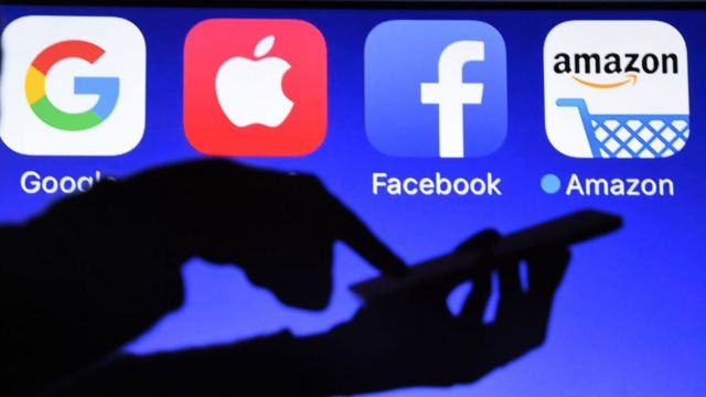 Sombra de mão mexendo no celular com tela maior atrás mostrando ícones do Google, Apple, Facebook e Amazon
